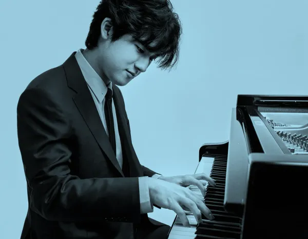 Yunchan Lim in a suit playing a grand piano with focus and concentration against a plain background.