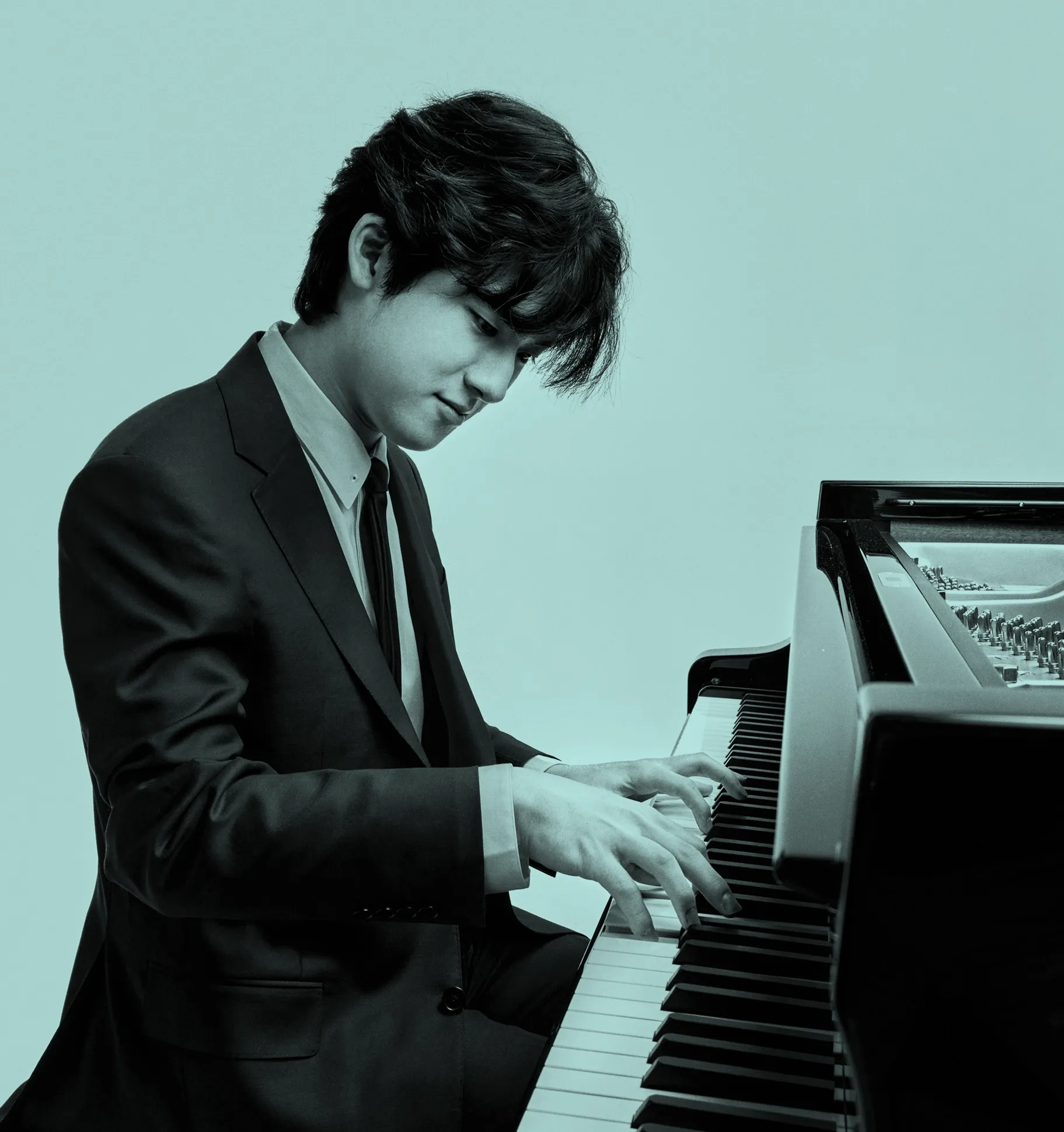 Yunchan Lim in a suit playing a grand piano with focus and concentration against a plain background.