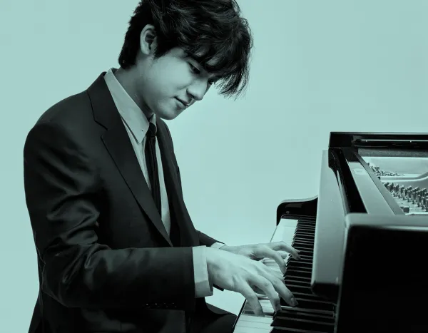 Yunchan Lim in a suit playing a grand piano with focus and concentration against a plain background.