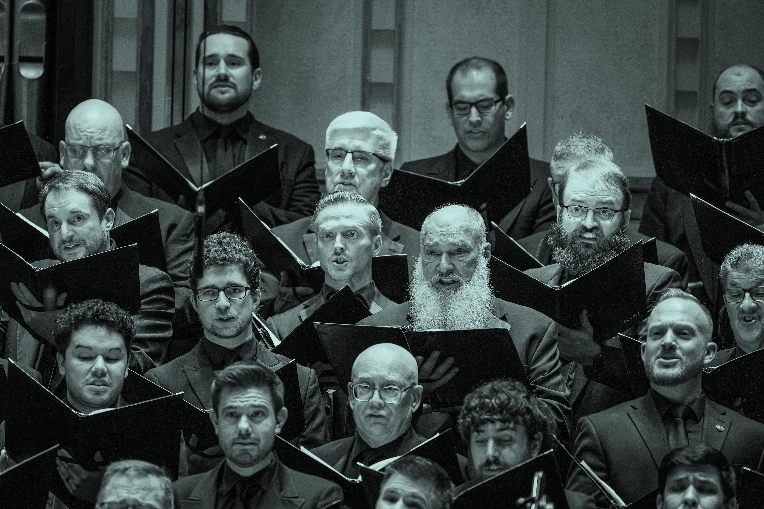 A men's choir performs, holding sheet music and wearing dark suits, with expressions of focus and intensity.