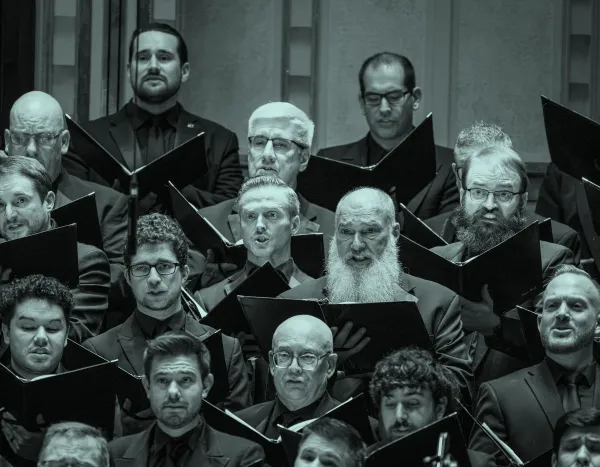 A men's choir performs, holding sheet music and wearing dark suits, with expressions of focus and intensity.