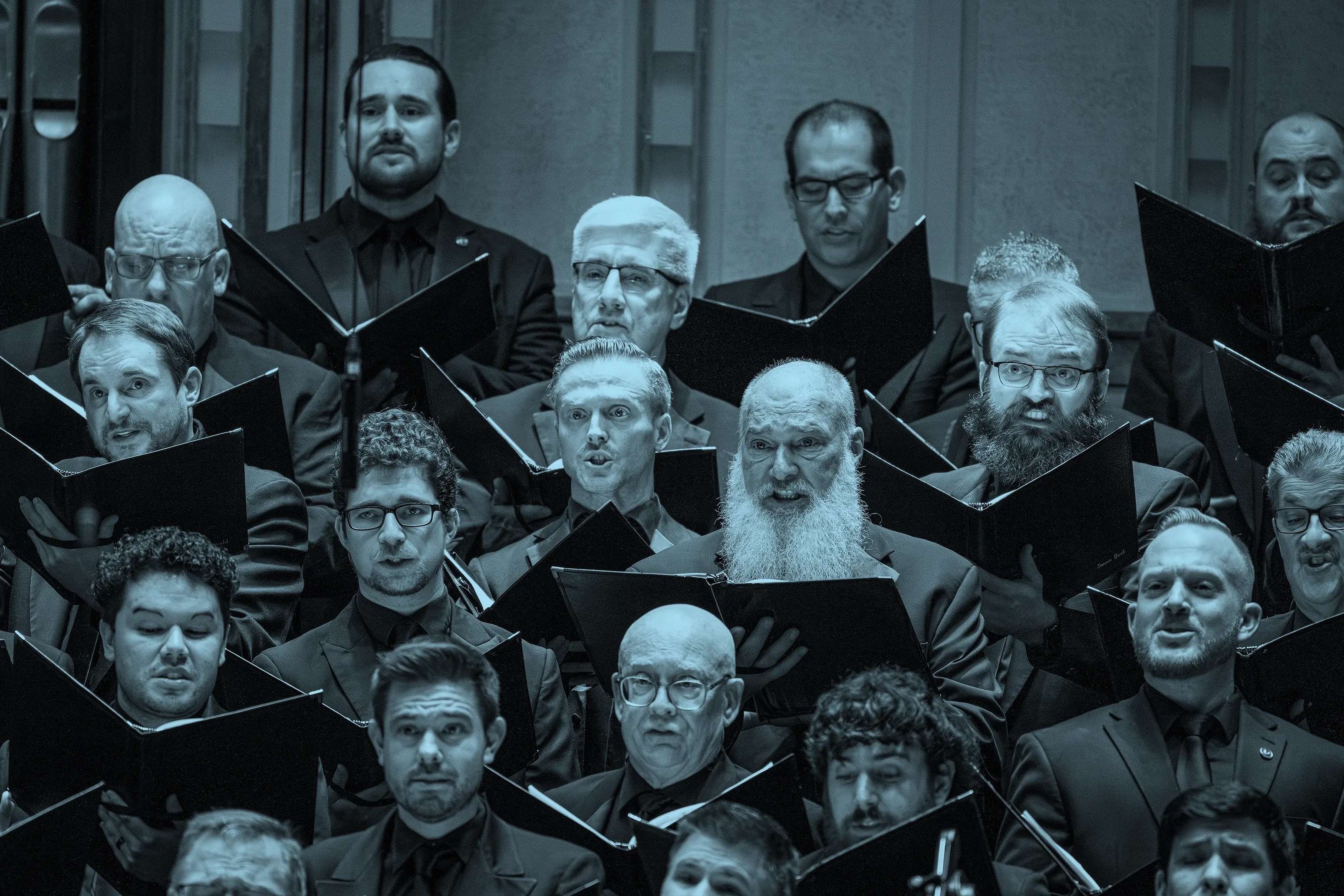 A men's choir performs, holding sheet music and wearing dark suits, with expressions of focus and intensity.