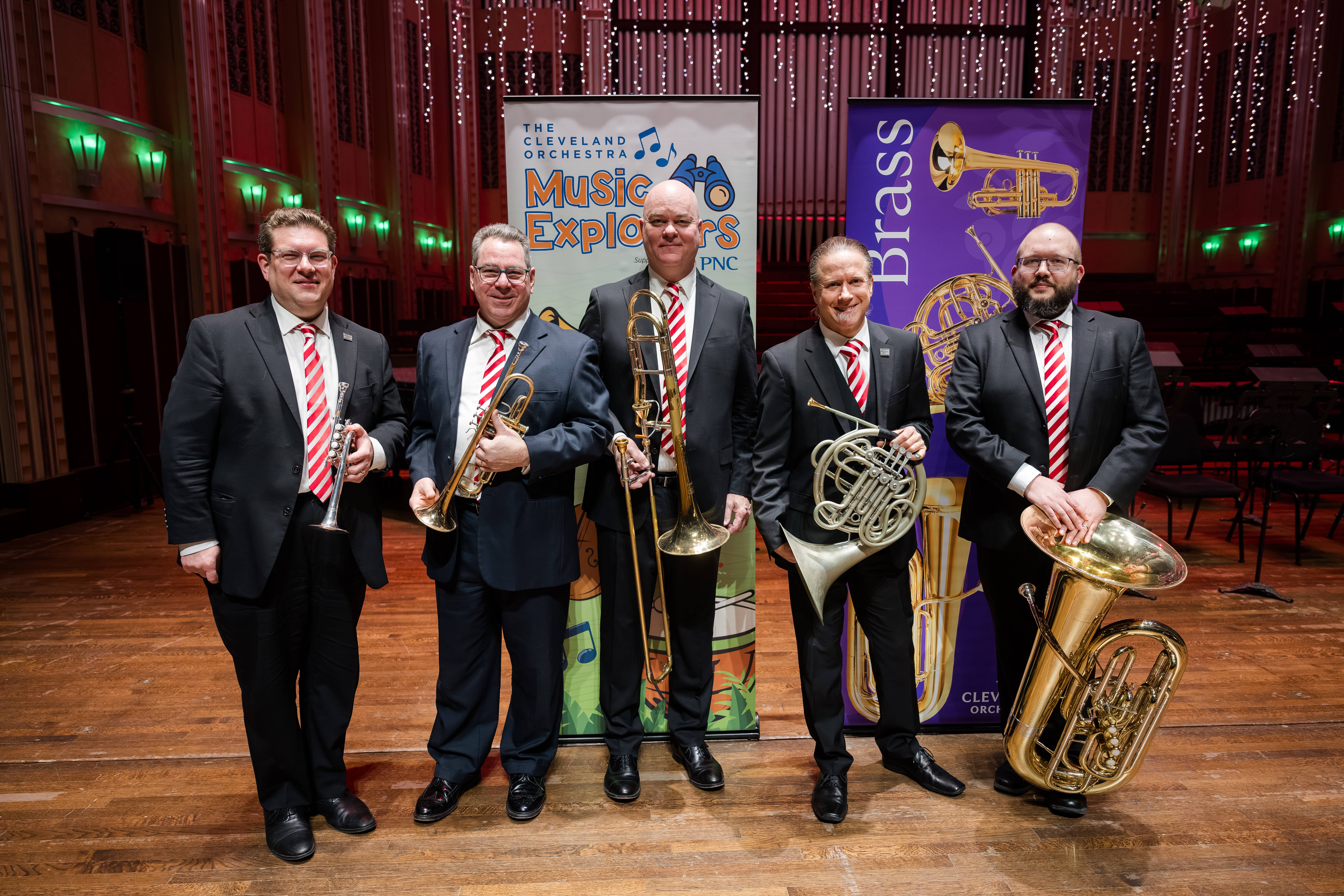 Five men in suits holding brass instruments in a theater lobby with concert banners and lit stage backdrop.