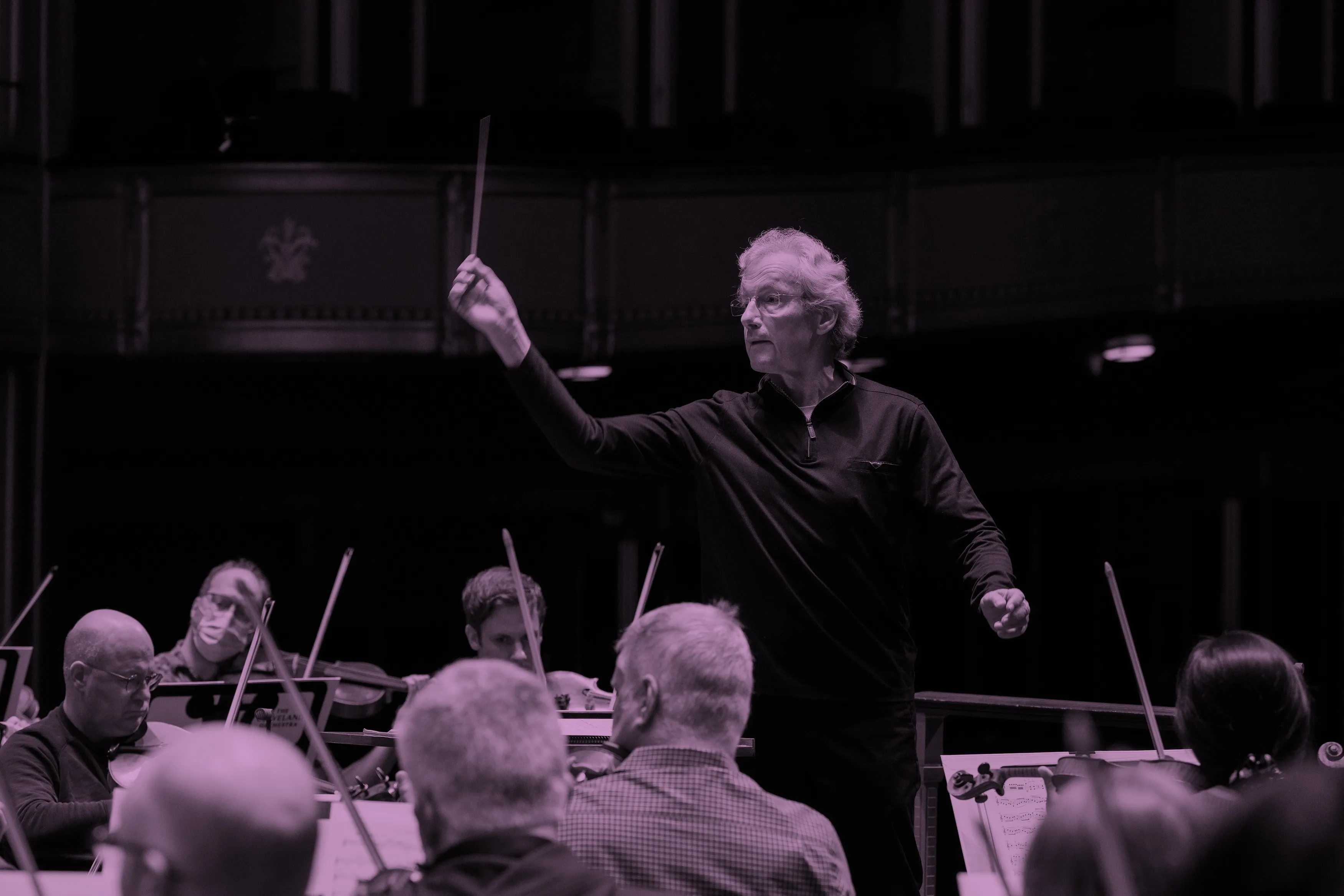 Franz Welser-Möst leading The Cleveland Orchestra with raised baton, musicians focused on their instruments in rehearsal.
