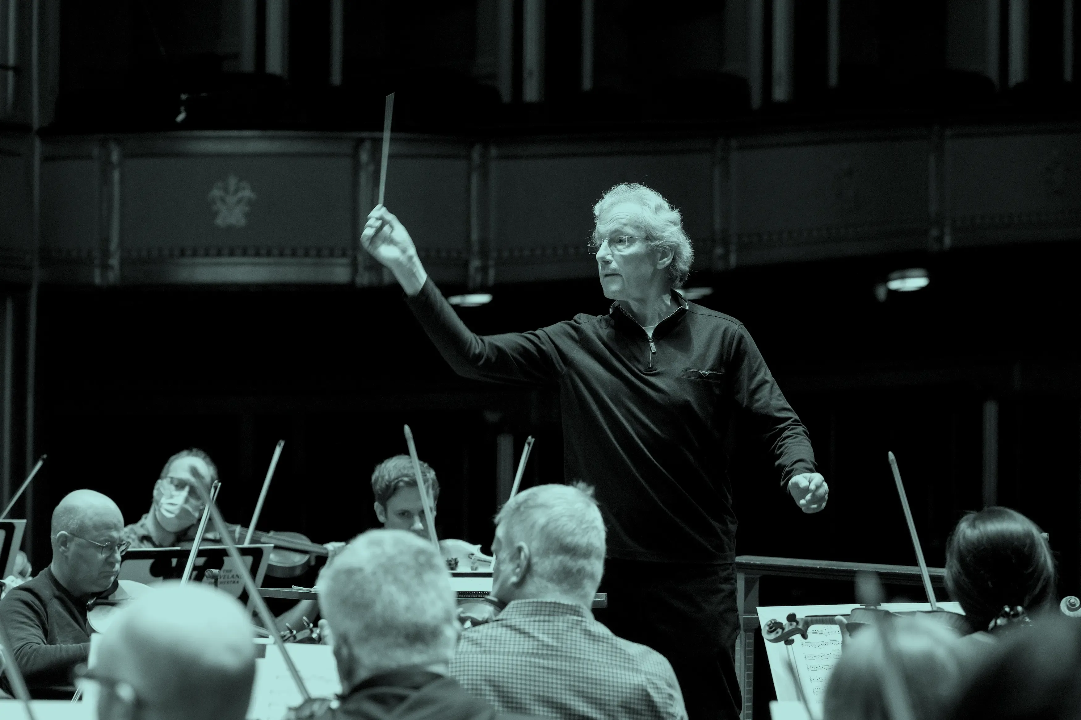 Franz Welser-Möst leading The Cleveland Orchestra with raised baton, musicians focused on their instruments in rehearsal.