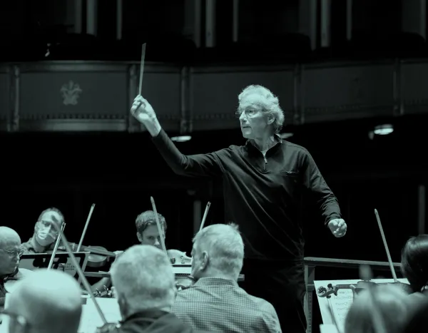 Franz Welser-Möst leading The Cleveland Orchestra with raised baton, musicians focused on their instruments in rehearsal.