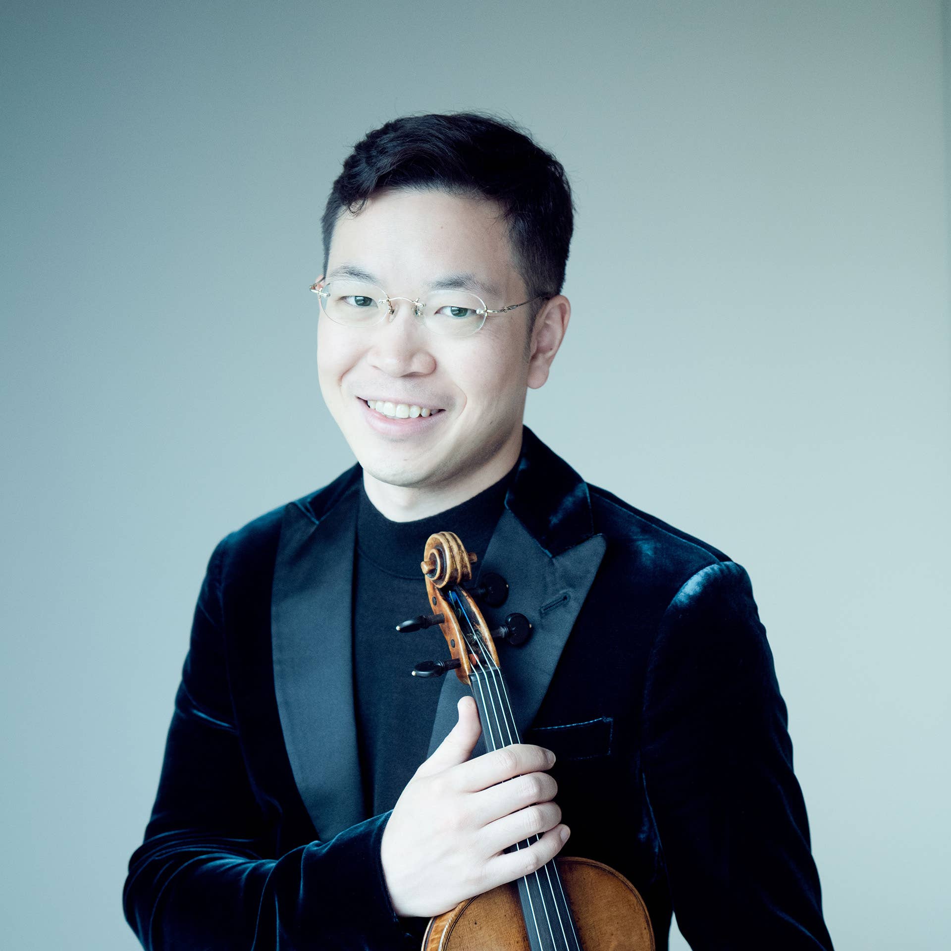 Paul Huang in a dark velvet jacket holding a violin against a light background.