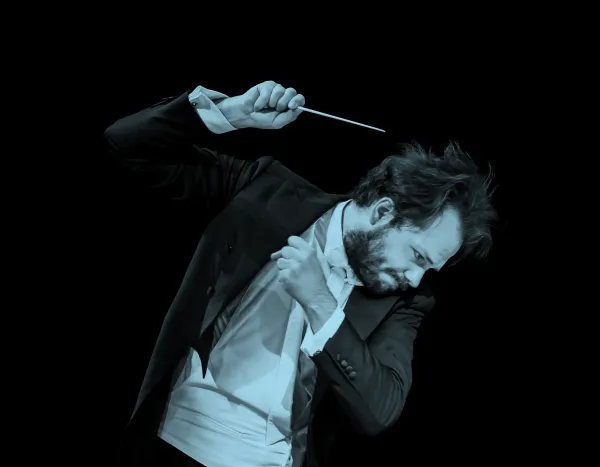 Conductor, Petr Popelka, passionately leading an orchestra, holding a baton, wearing a tuxedo, against a black background.