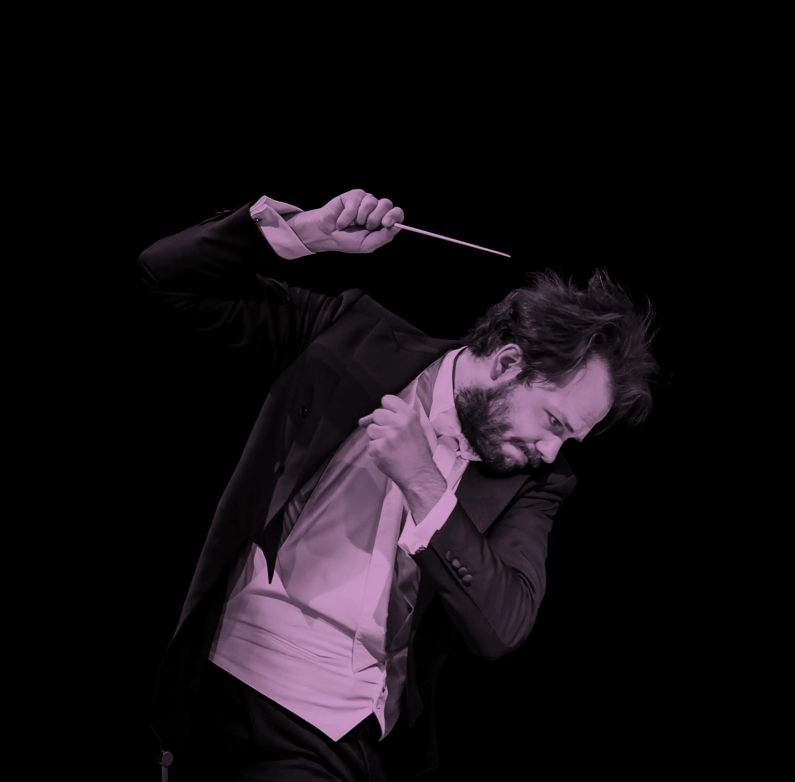 Conductor, Petr Popelka, passionately leading an orchestra, holding a baton, wearing a tuxedo, against a black background.