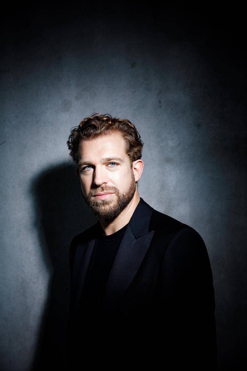 Julian Prégardien wearing a black suit, standing against a dark, textured background with dramatic lighting.