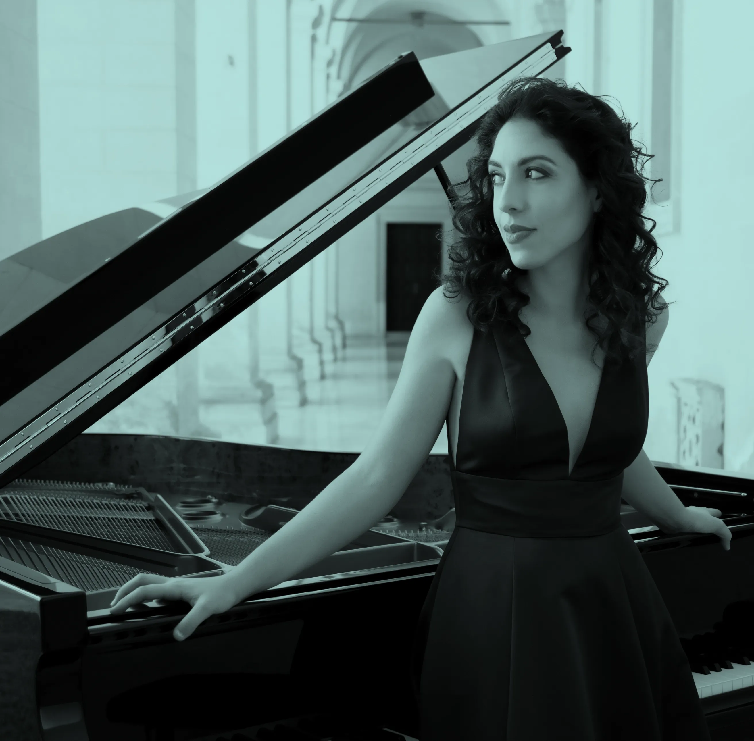 Beatrice Rana in a dark blue dress stands by a grand piano in an elegant hallway, looking to the side with a thoughtful expression.