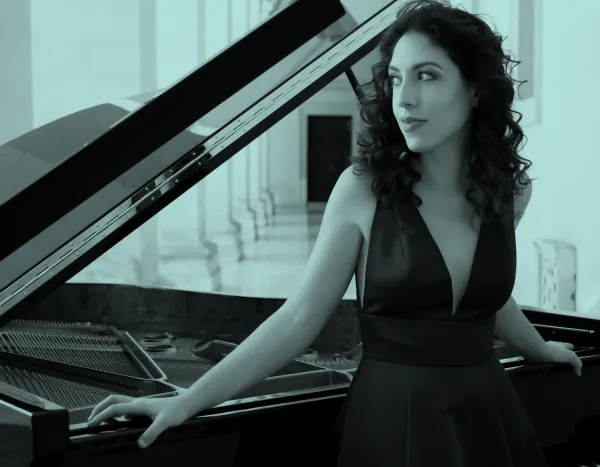 Beatrice Rana in a dark blue dress stands by a grand piano in an elegant hallway, looking to the side with a thoughtful expression.