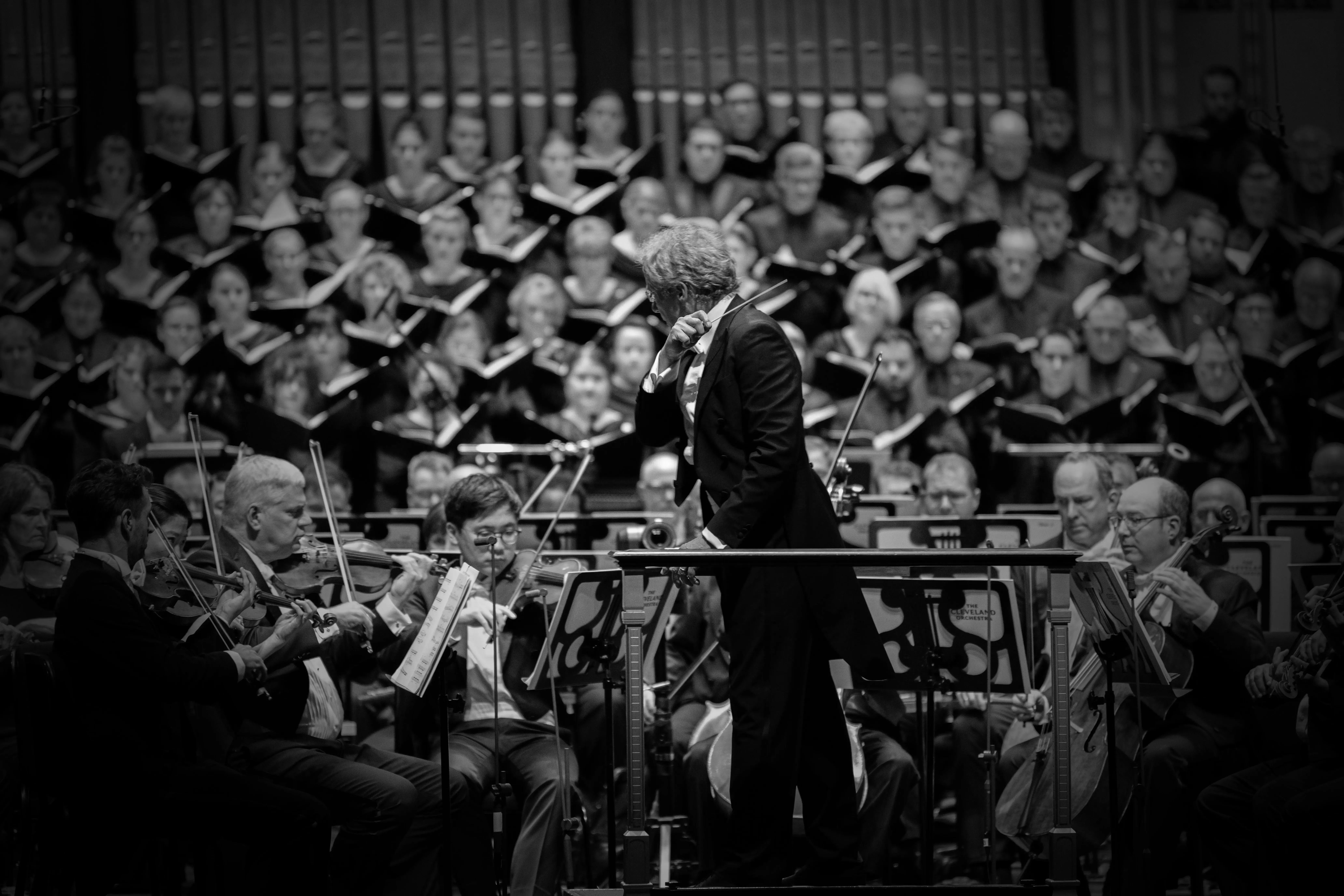 Conductor in black suit leads orchestra in monochrome photo with musicians holding violins and instruments.