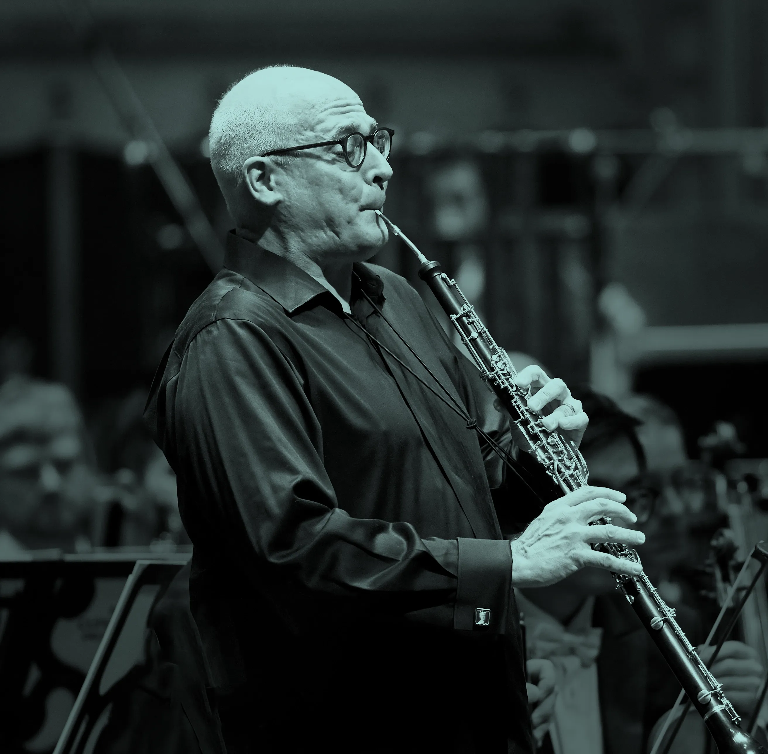 Robert Walters playing an oboe in an orchestra, wearing glasses and a dark shirt, with musicians blurred in the background.