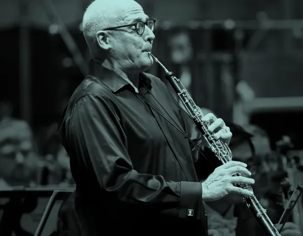 Robert Walters playing an oboe in an orchestra, wearing glasses and a dark shirt, with musicians blurred in the background.