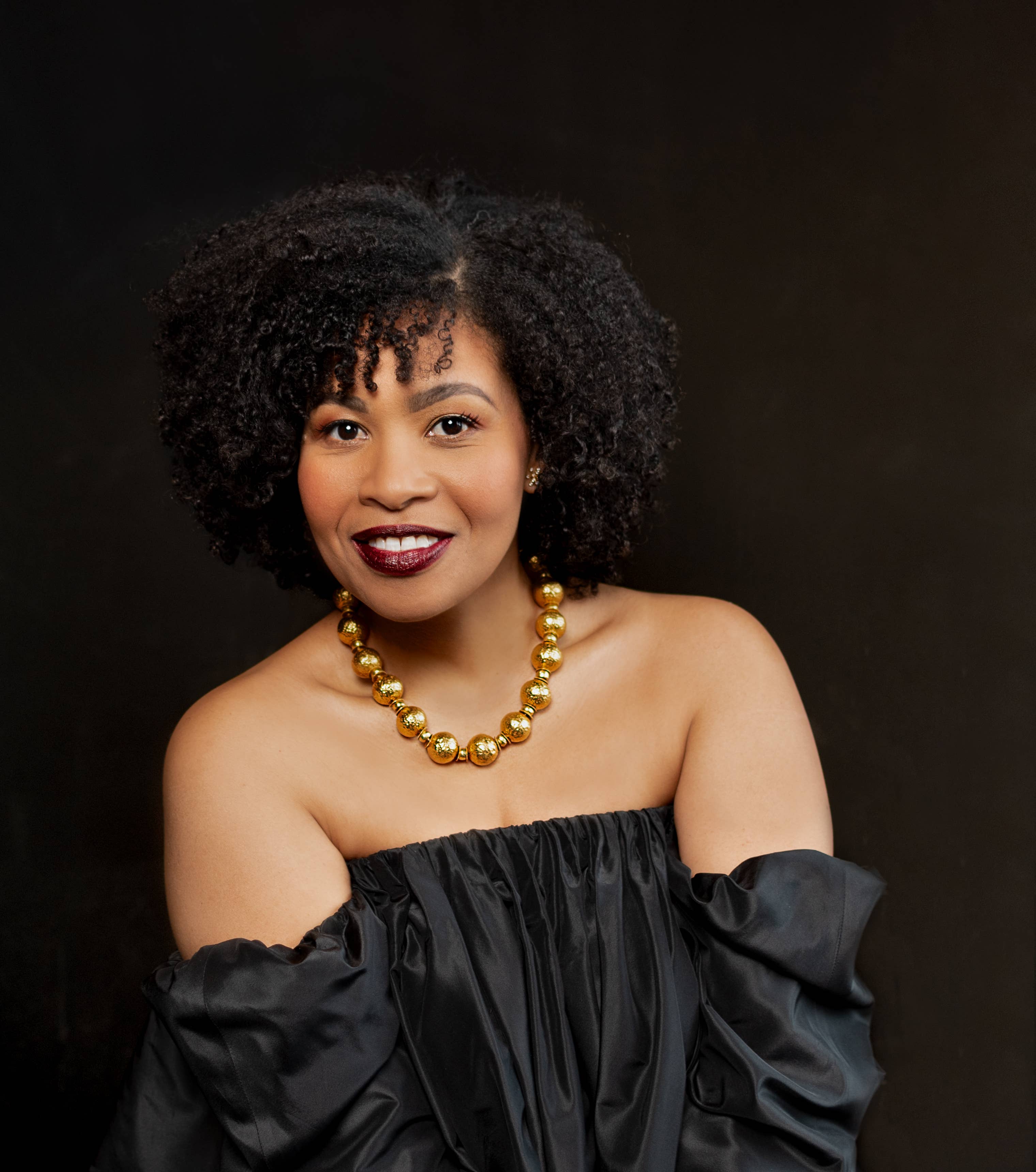 Golda Schultz posing for headshot in a black dress and gold necklace