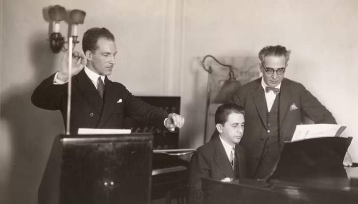 A man playing a theremin stands next to a man playing a piano, with a third man, in glasses, looking on