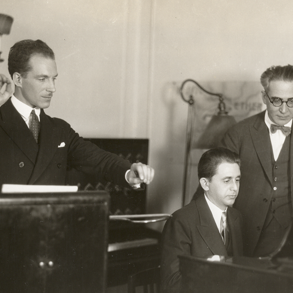 A man playing a theremin stands next to a man playing a piano, with a third man, in glasses, looking on