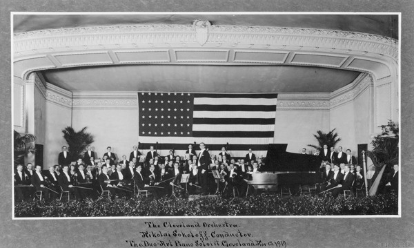 An orchestra sits on stage with a piano and a large America flag.
