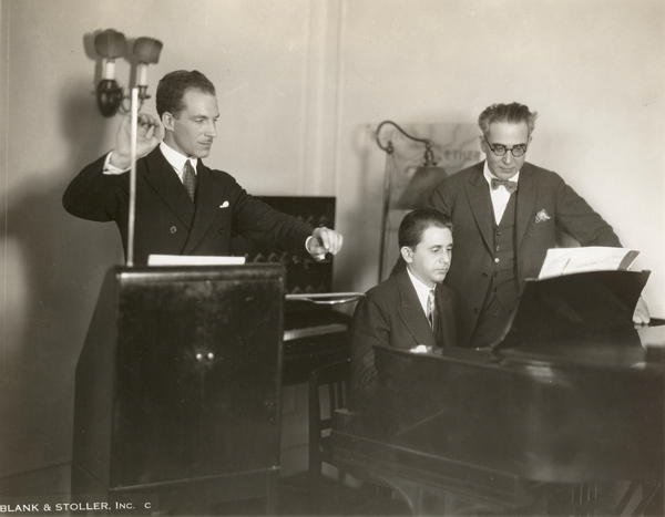 A man playing a theremin stands next to a man playing a piano, with a third man, in glasses, looking on