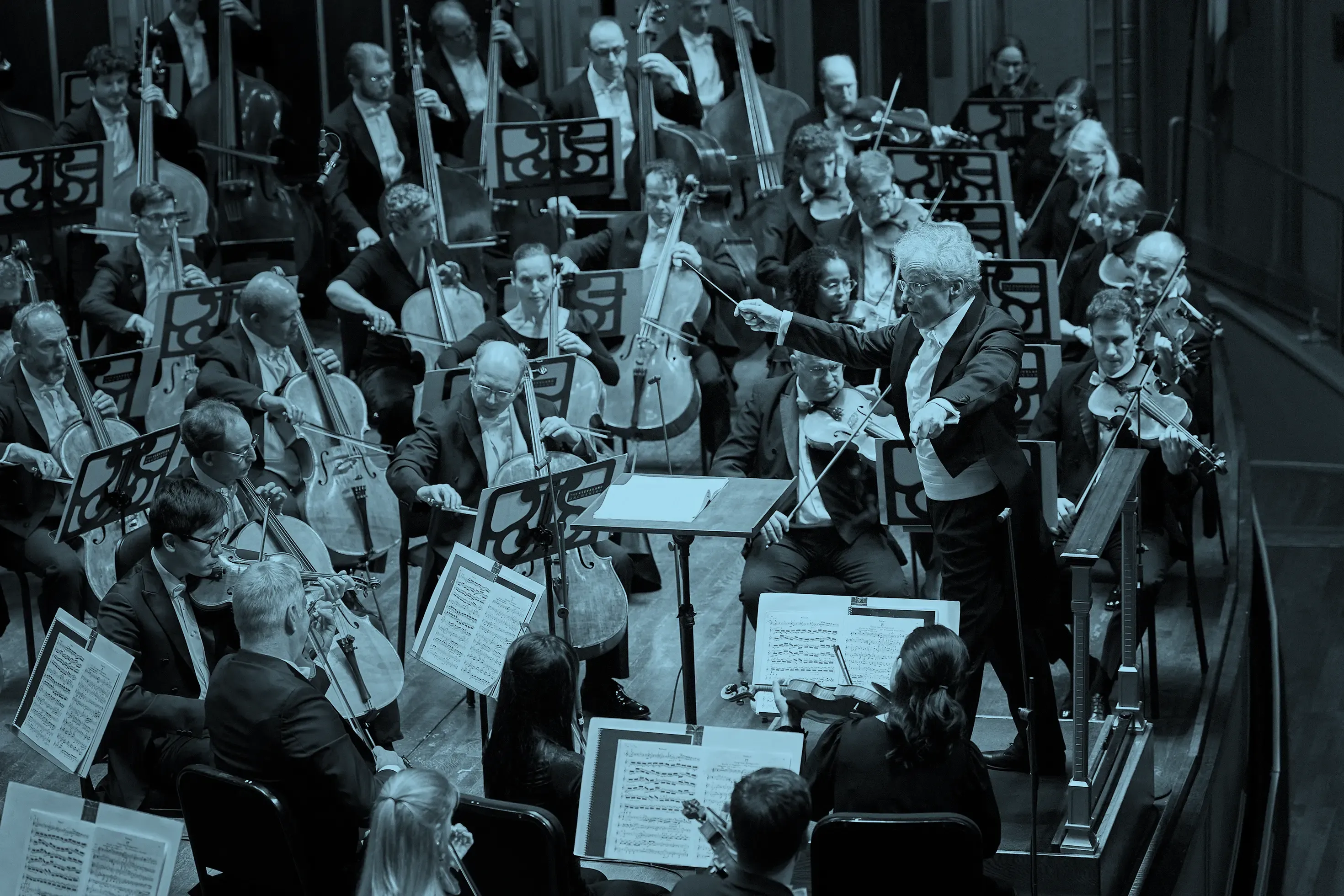 Franz Welser Möst in a tuxedo energetically leads an orchestra with a raised baton, surrounded by musicians playing string instruments.