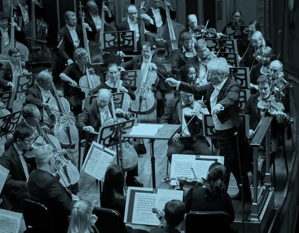 Franz Welser Möst in a tuxedo energetically leads an orchestra with a raised baton, surrounded by musicians playing string instruments.