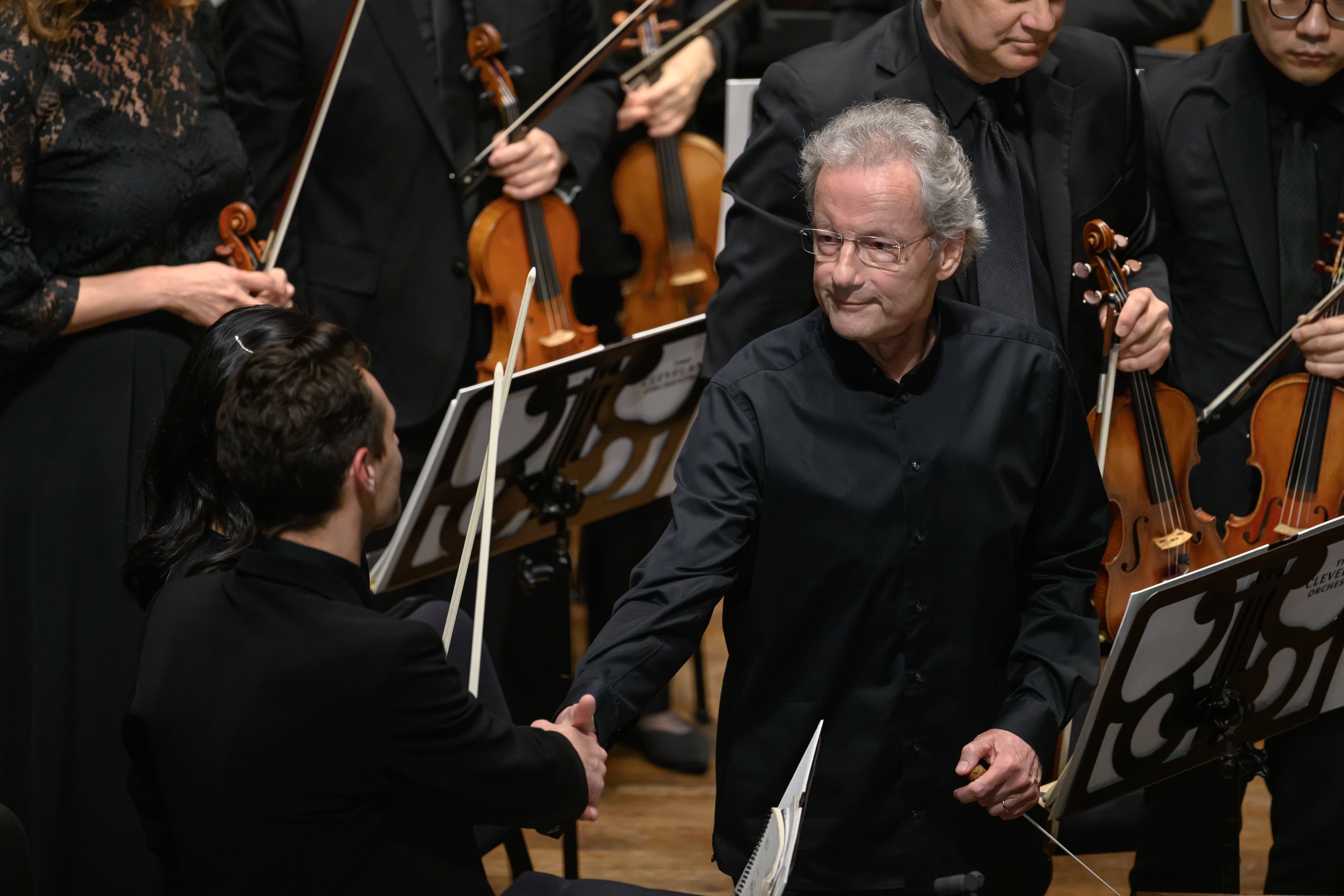 Orchestra conductor with baton, while string musicians play violins behind him during performance.