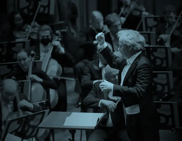 Franz Welser Möst leading The Cleveland Orchestra with musicians playing string instruments, including cellos and violins, in a dimly lit setting.