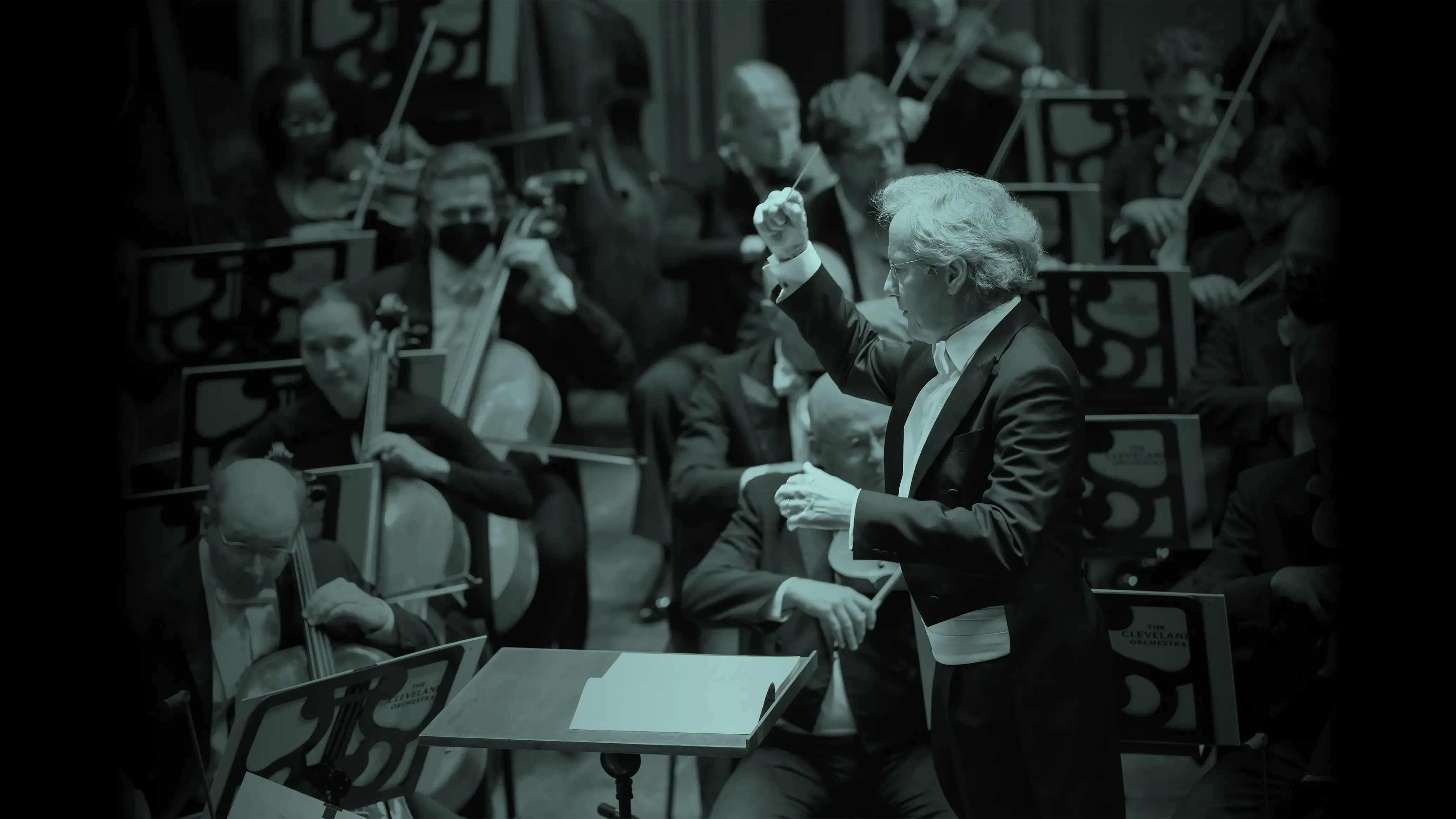 Franz Welser Möst leading The Cleveland Orchestra with musicians playing string instruments, including cellos and violins, in a dimly lit setting.