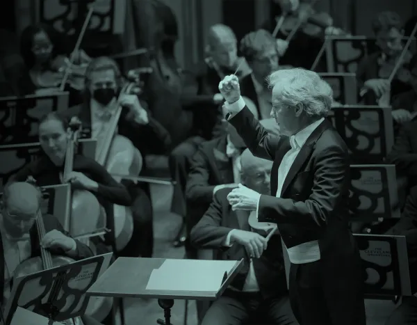 Franz Welser Möst leading The Cleveland Orchestra with musicians playing string instruments, including cellos and violins, in a dimly lit setting.