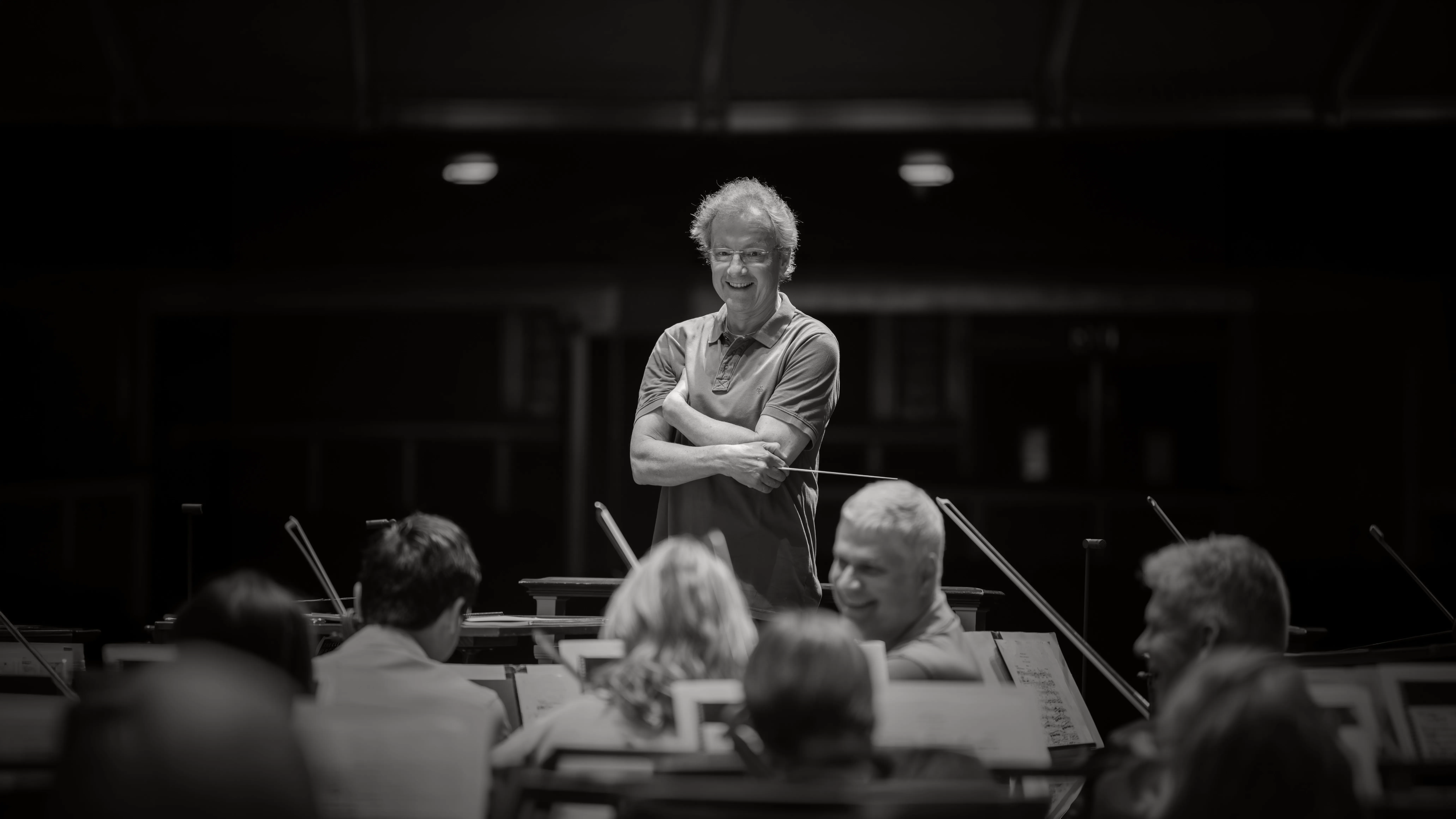 Smiling conductor in a dark concert hall, holding a baton while leading an orchestra in front of music stands