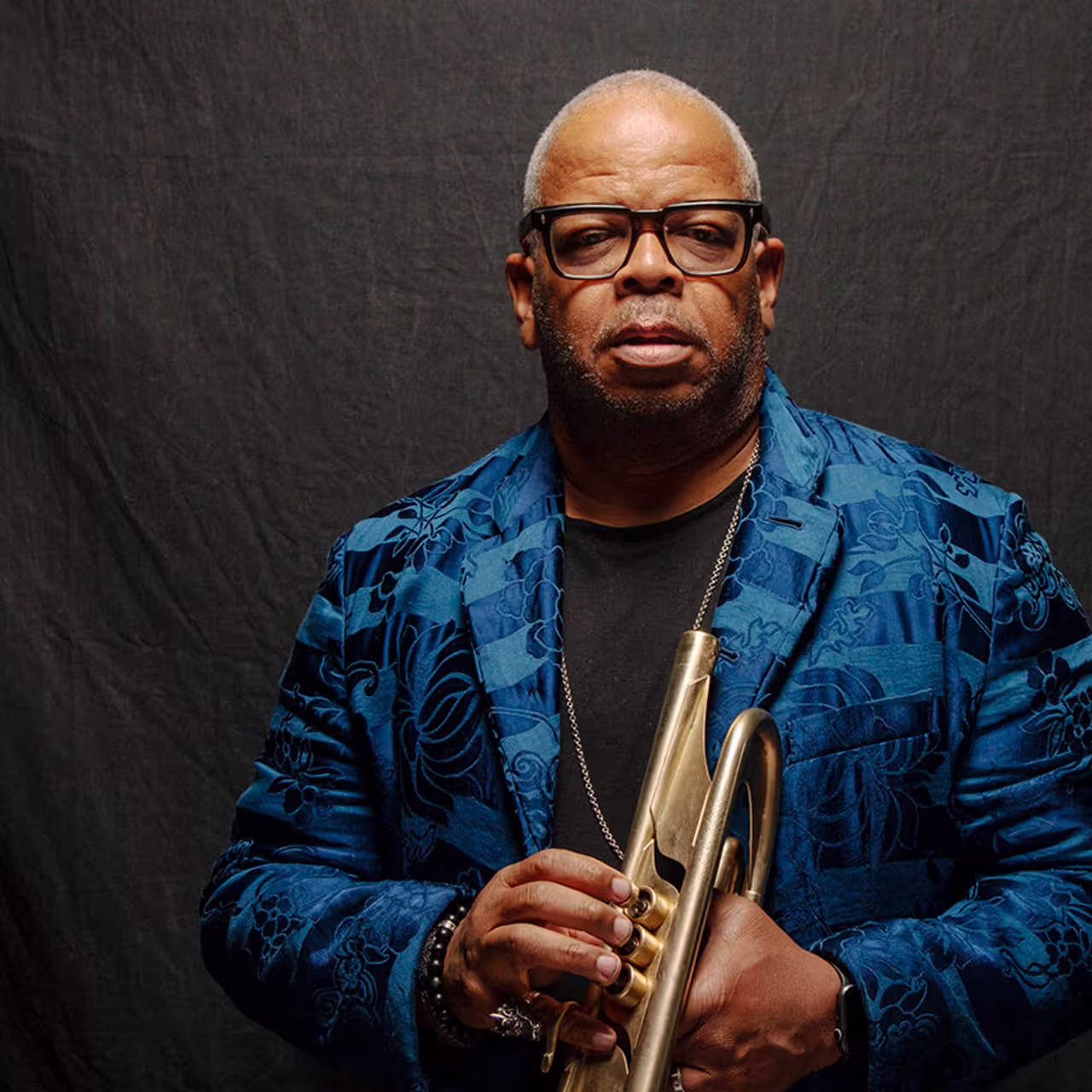 Terence Blanchard in a blue jacket holding a trumpet in font of a dark gray backdrop
