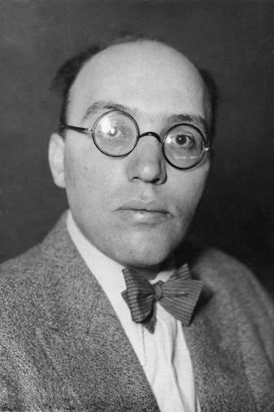 Kurt Weill with round glasses and bow tie, wearing a tweed jacket, looking directly at the camera against a dark background.