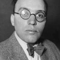 Kurt Weill with round glasses and bow tie, wearing a tweed jacket, looking directly at the camera against a dark background.