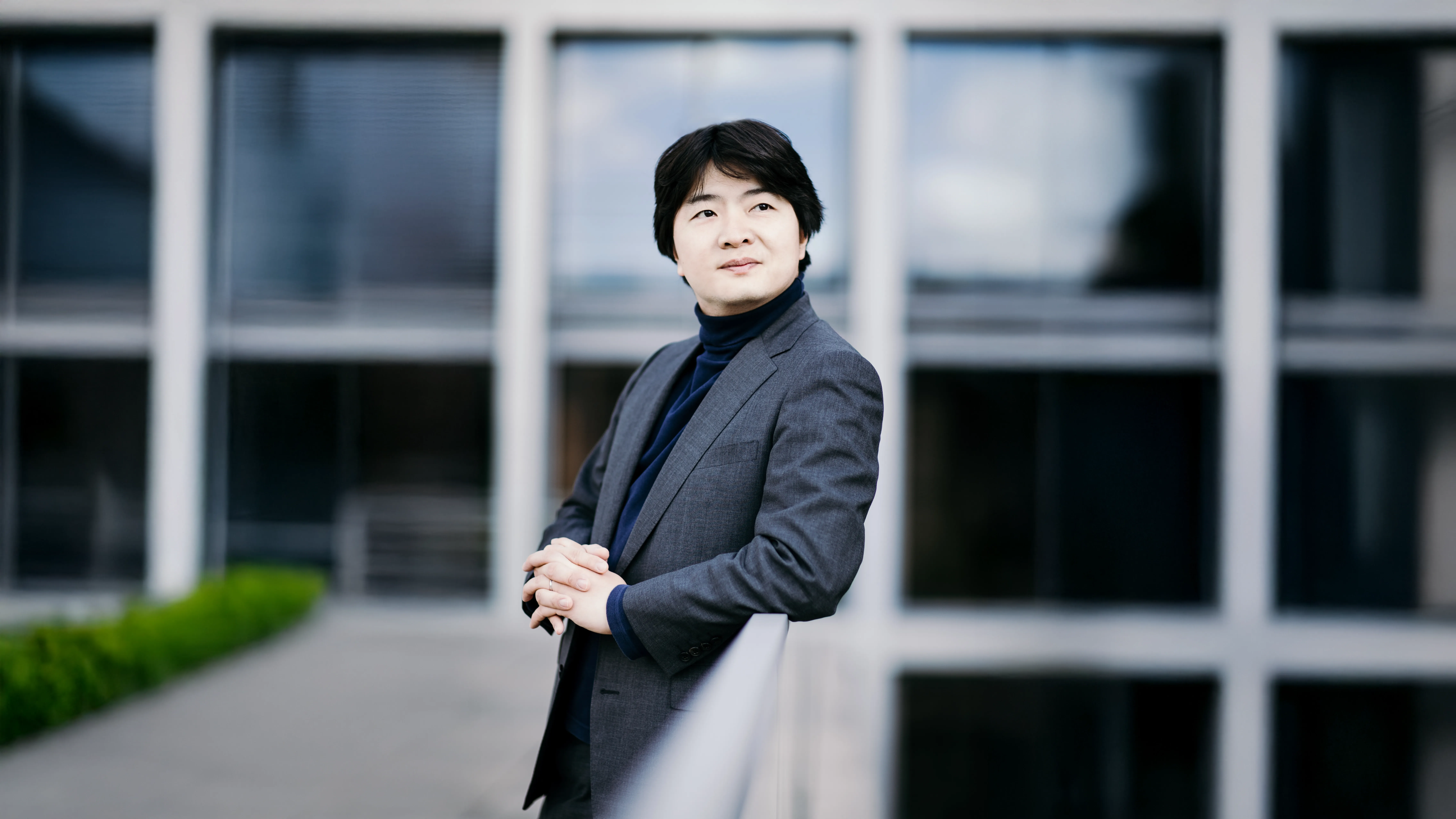 Kazuki Yamada in a gray suit and dark turtleneck leans on a railing, looking thoughtfully into the distance, with a building behind.