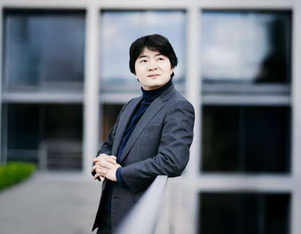Kazuki Yamada in a gray suit and dark turtleneck leans on a railing, looking thoughtfully into the distance, with a building behind.