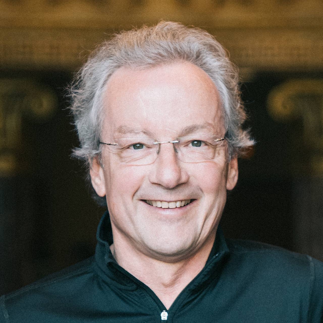 A close-up portrait of Franz Welser-Möst, a smiling middle-aged man with light gray hair and glasses. He is dressed in a black jacket and has a friendly, approachable expression. The background is blurred, with hints of architectural elements.