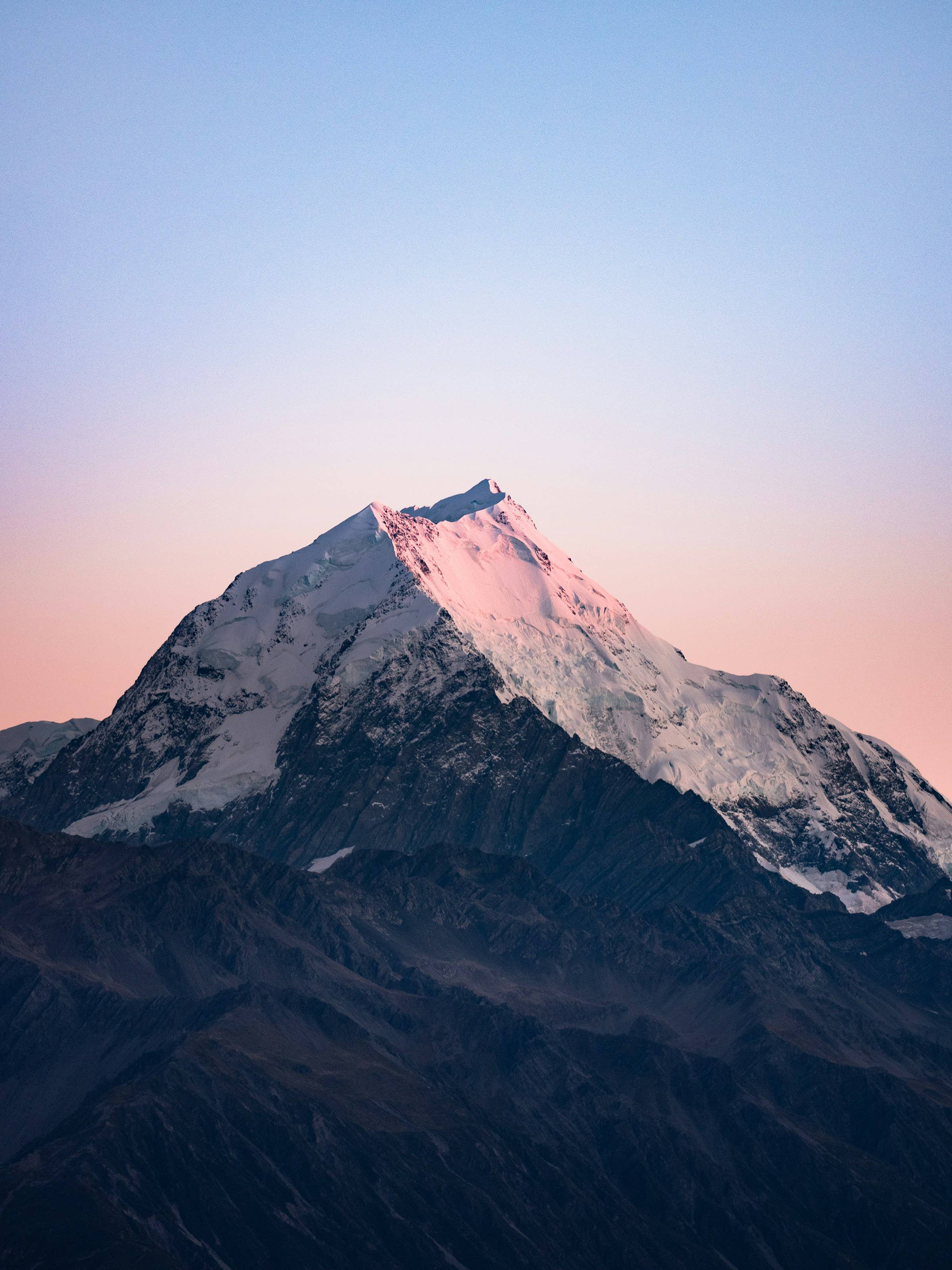Snowcapped mountain peak at sunset, with pastel pink sky and dark rocky slopes below