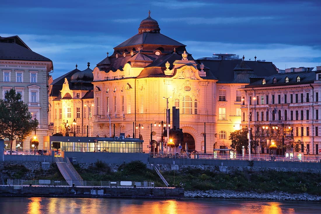 Bratislava, Slovakia, exterior of Reduta Hall Historic building with ornate architecture illuminated at night, reflecting on a river under a deep blue sky.