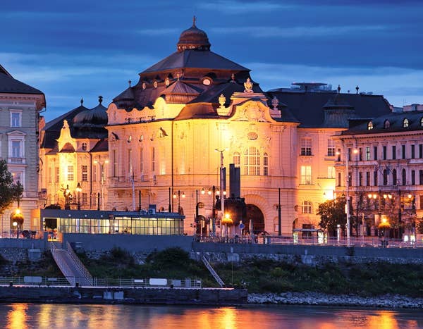 Bratislava, Slovakia, exterior of Reduta Hall Historic building with ornate architecture illuminated at night, reflecting on a river under a deep blue sky.