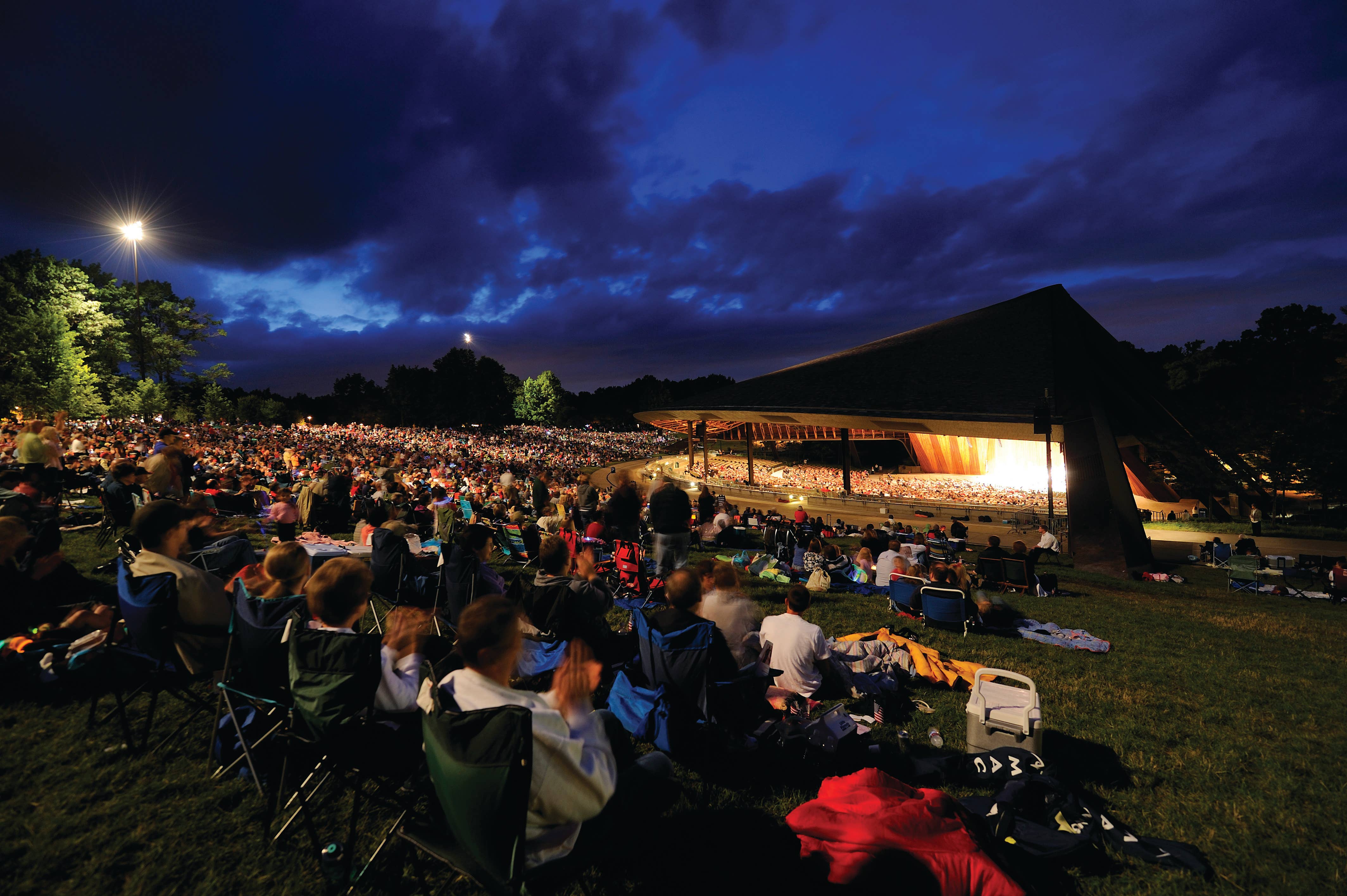 under a dark blue sky, people in lawn chairs look forward to an illuminated stage