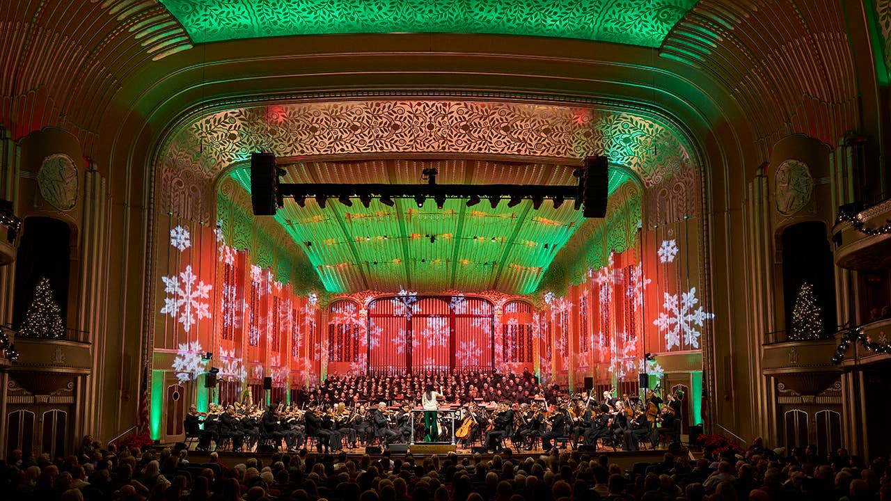 An orchestra sits on a stage bathed in green and red light with snowflake projections