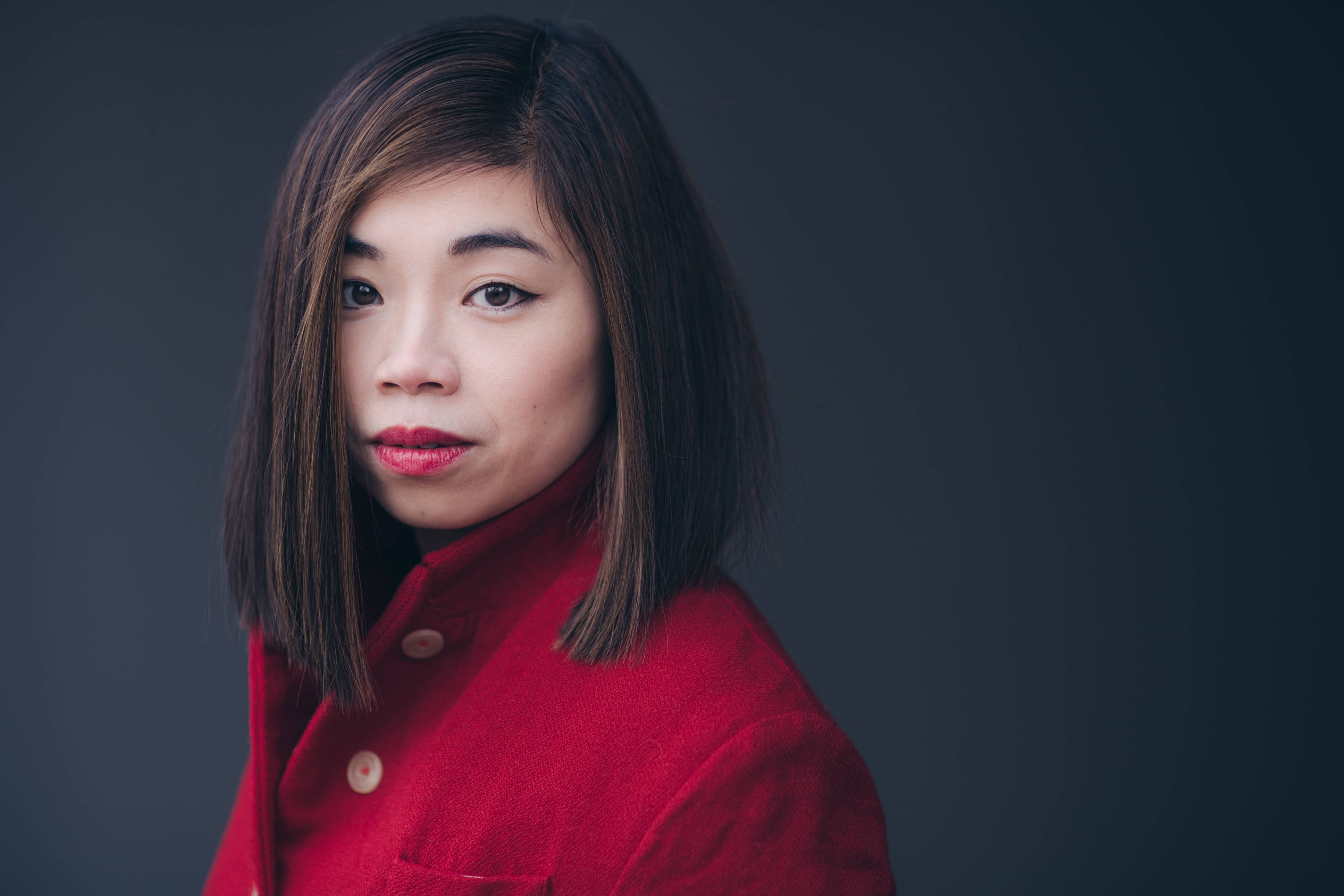 Elim Chan with straight brown hair wearing a red coat, looking at the camera against a dark background.