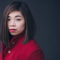 Elim Chan with straight brown hair wearing a red coat, looking at the camera against a dark background.