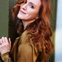 Sara Jakubiak with wavy red hair and a brown blouse leans against a green wall, smiling softly at the camera.