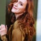 Sara Jakubiak with wavy red hair and a brown blouse leans against a green wall, smiling softly at the camera.
