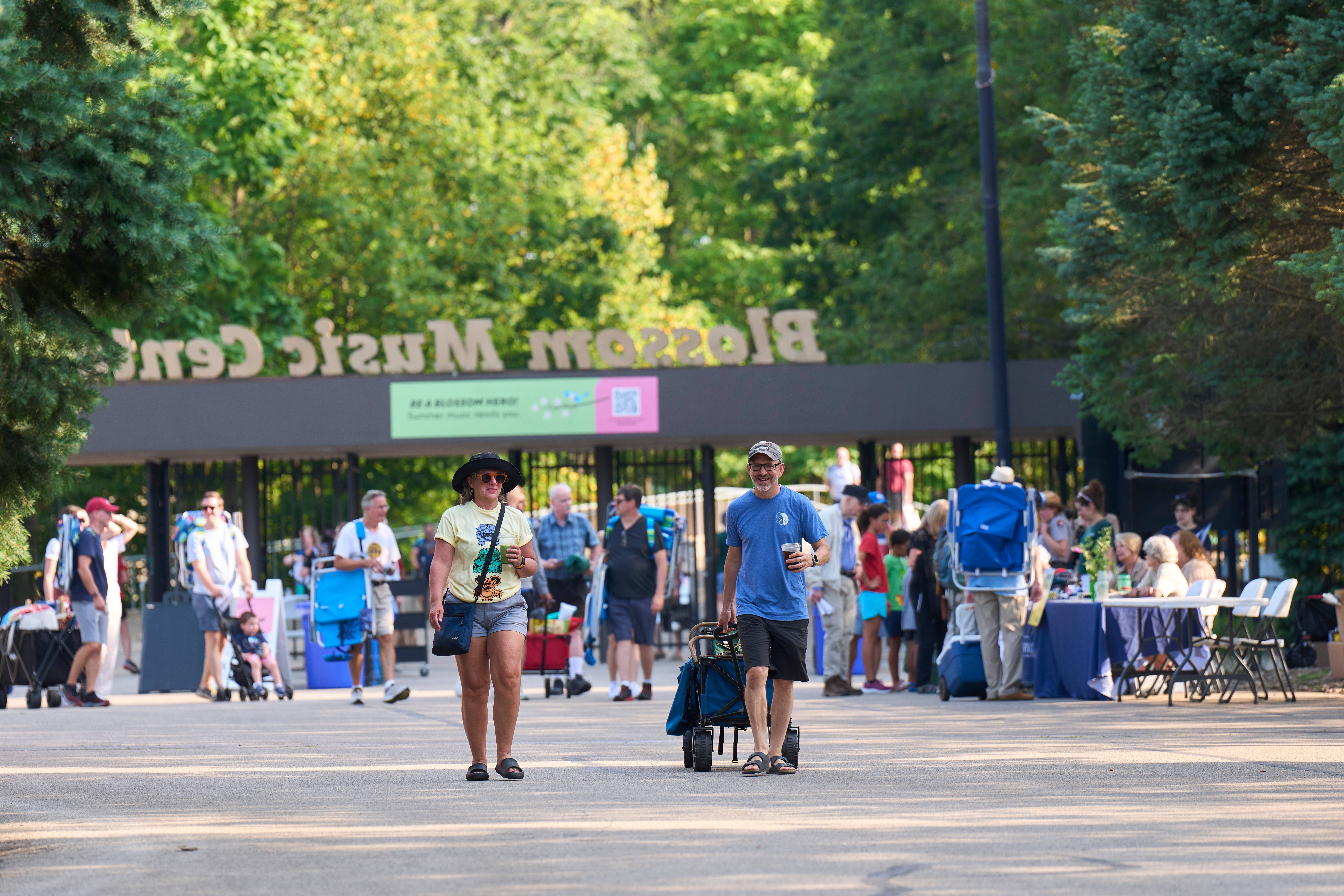 People walking through Blossom Music Center gate entrance, carrying coolers and drinks, surrounded by trees and a crowd.