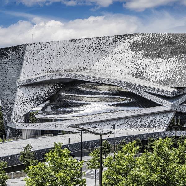 Modern architectural building with a metallic, angular facade under a partly cloudy sky, surrounded by green trees.