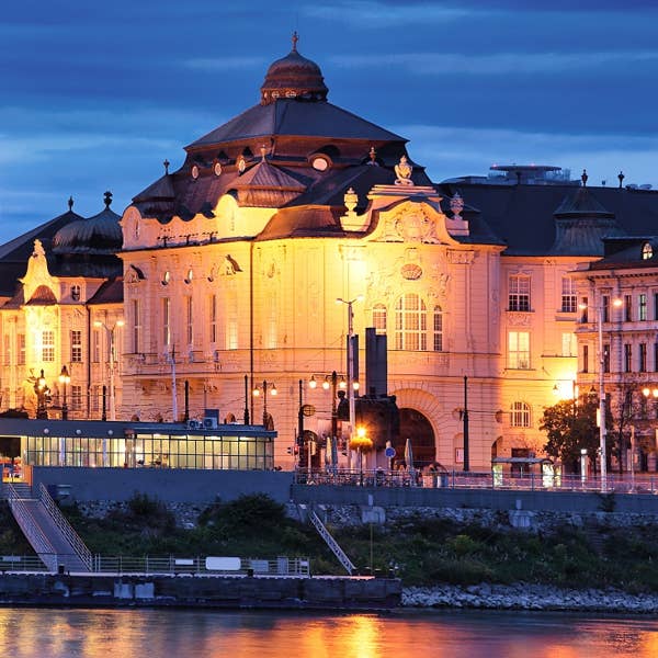 Bratislava, Slovakia, exterior of Reduta Hall Historic building with ornate architecture illuminated at night, reflecting on a river under a deep blue sky.