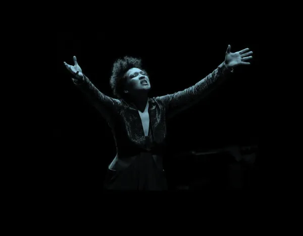 Julia Bullock with arms raised, passionately performing on stage in dramatic lighting.